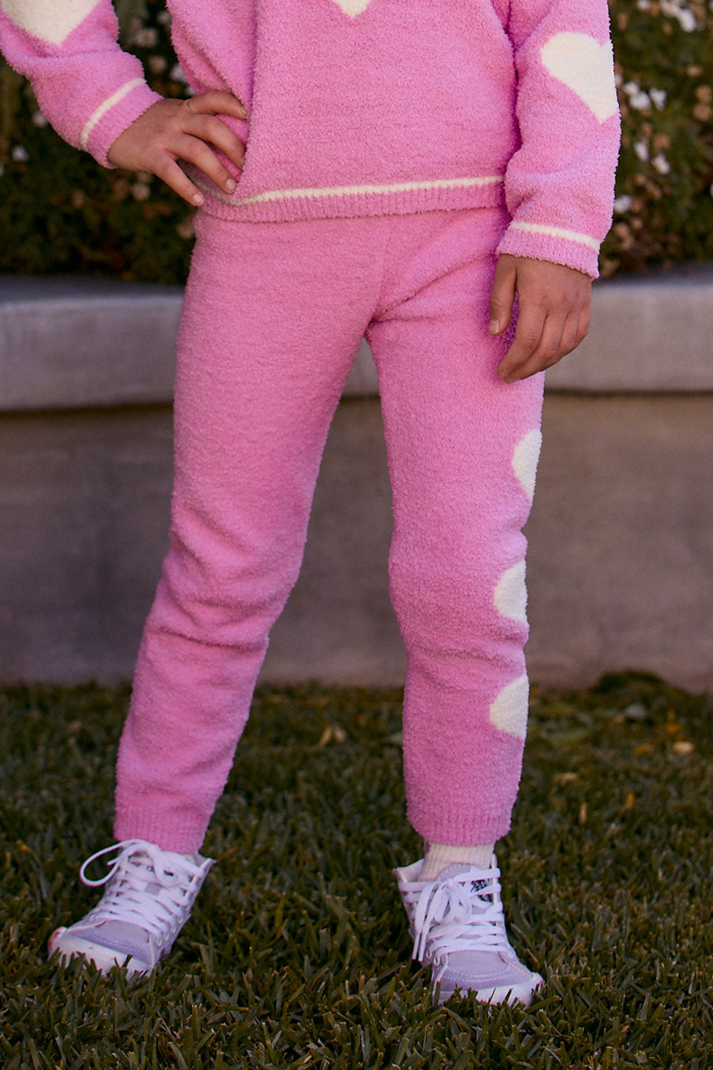Chaser Kids Holiday Hug Girls Pants in pink with white hearts down one leg, front view outdoors beside a low wall and greenery