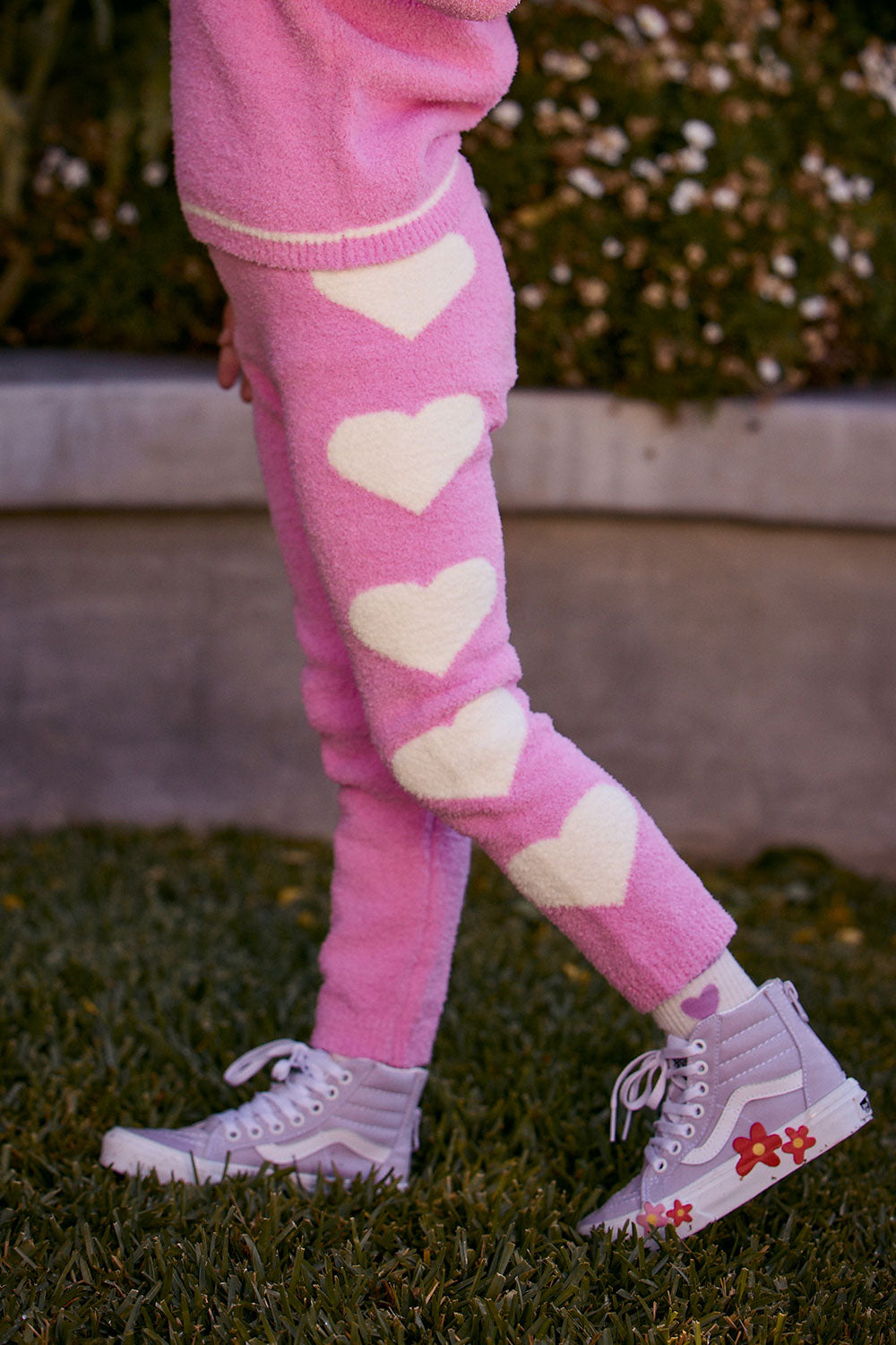 Chaser Kids Holiday Hug Girls Pants in pink, side detail of white hearts outdoors beside a low wall and greenery