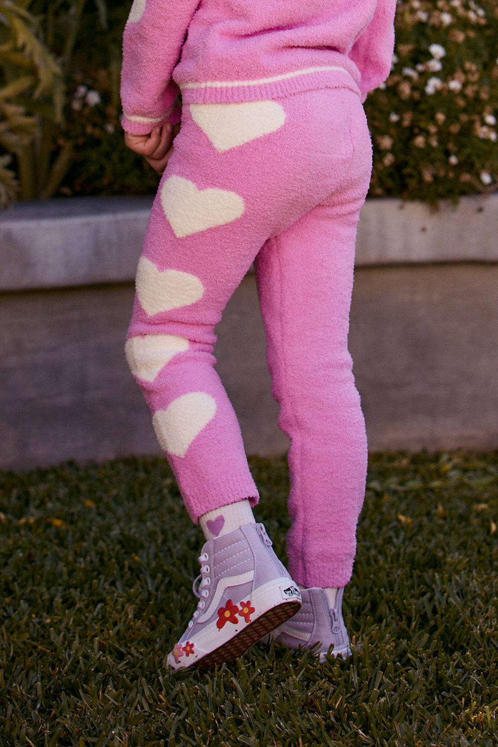 Chaser Kids Holiday Hug Girls Pants in pink with white hearts, rear detail outdoors beside a low wall and greenery