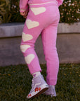 Chaser Kids Holiday Hug Girls Pants in pink with white hearts, rear detail outdoors beside a low wall and greenery