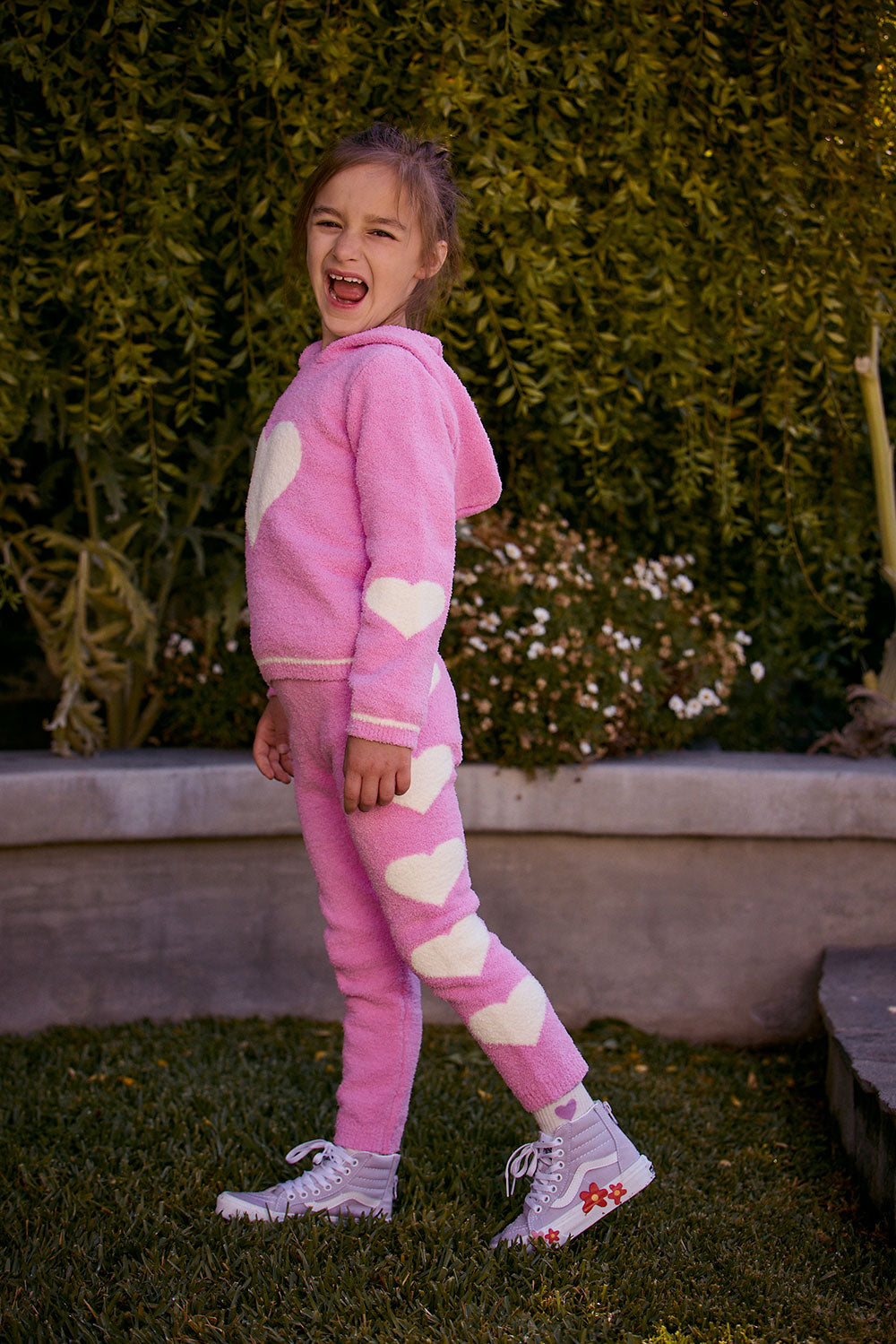 Chaser Kids Holiday Hug Girls Pants in pink with white hearts, full-body side view outdoors beside a low wall and greenery