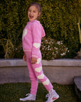 Chaser Kids Holiday Hug Girls Pants in pink with white hearts, full-body side view outdoors beside a low wall and greenery