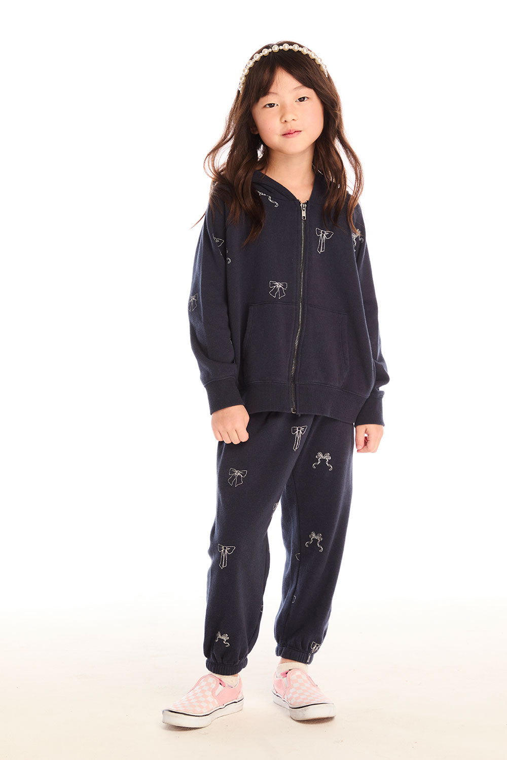 Full body view of girl wearing Chaser’s navy blue Tossed Glitter Bows Pants with embroidered bows and matching zip up hoodie on a white background