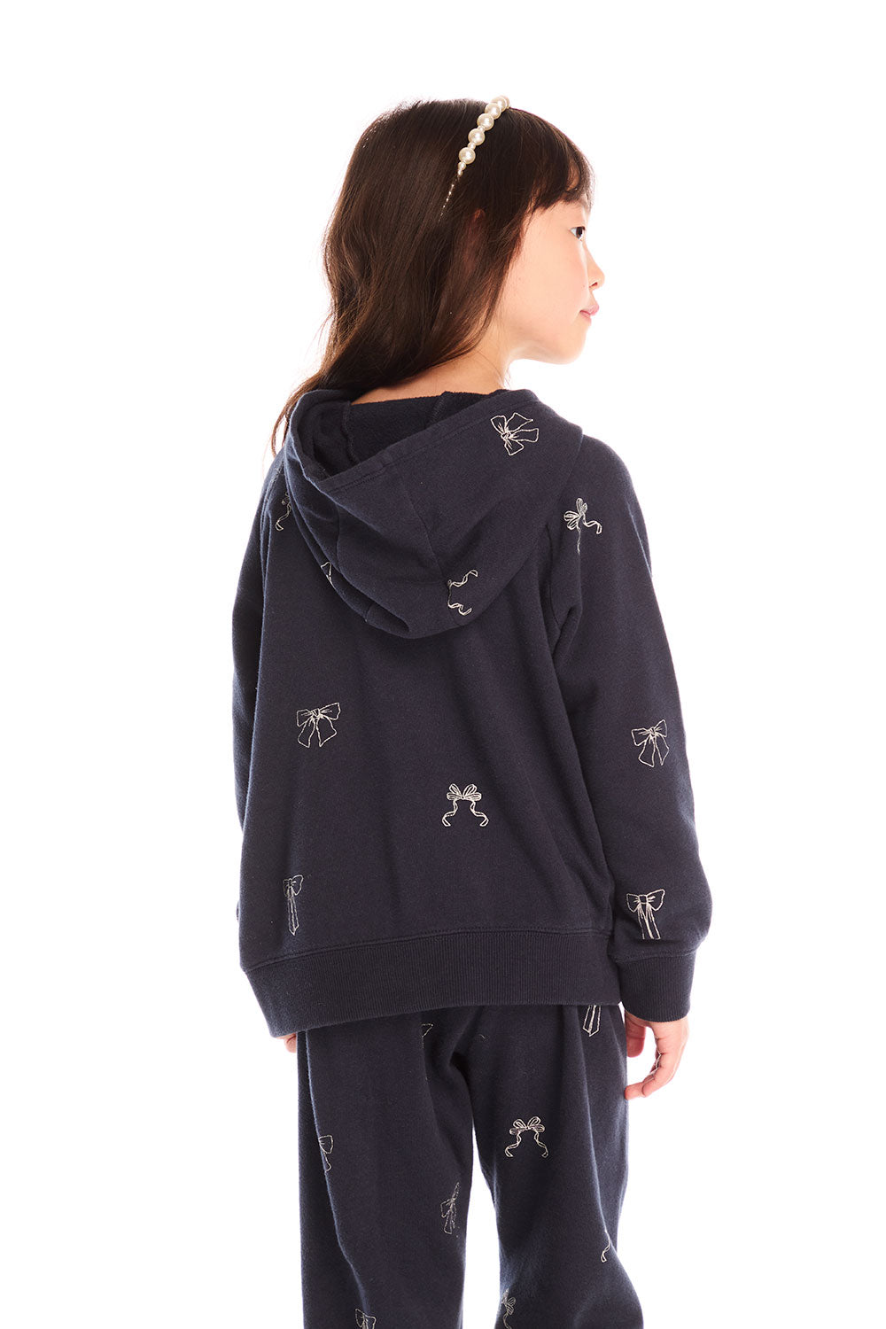 Back view of girl wearing Chaser’s navy blue Tossed Glitter Bows Zip Up Hoodie with embroidered bows on a white background