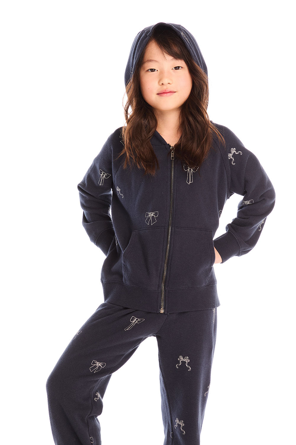 Girl wearing Chaser’s navy blue Tossed Glitter Bows Zip Up Hoodie with hood up and matching pants with embroidered bow designs on a white background