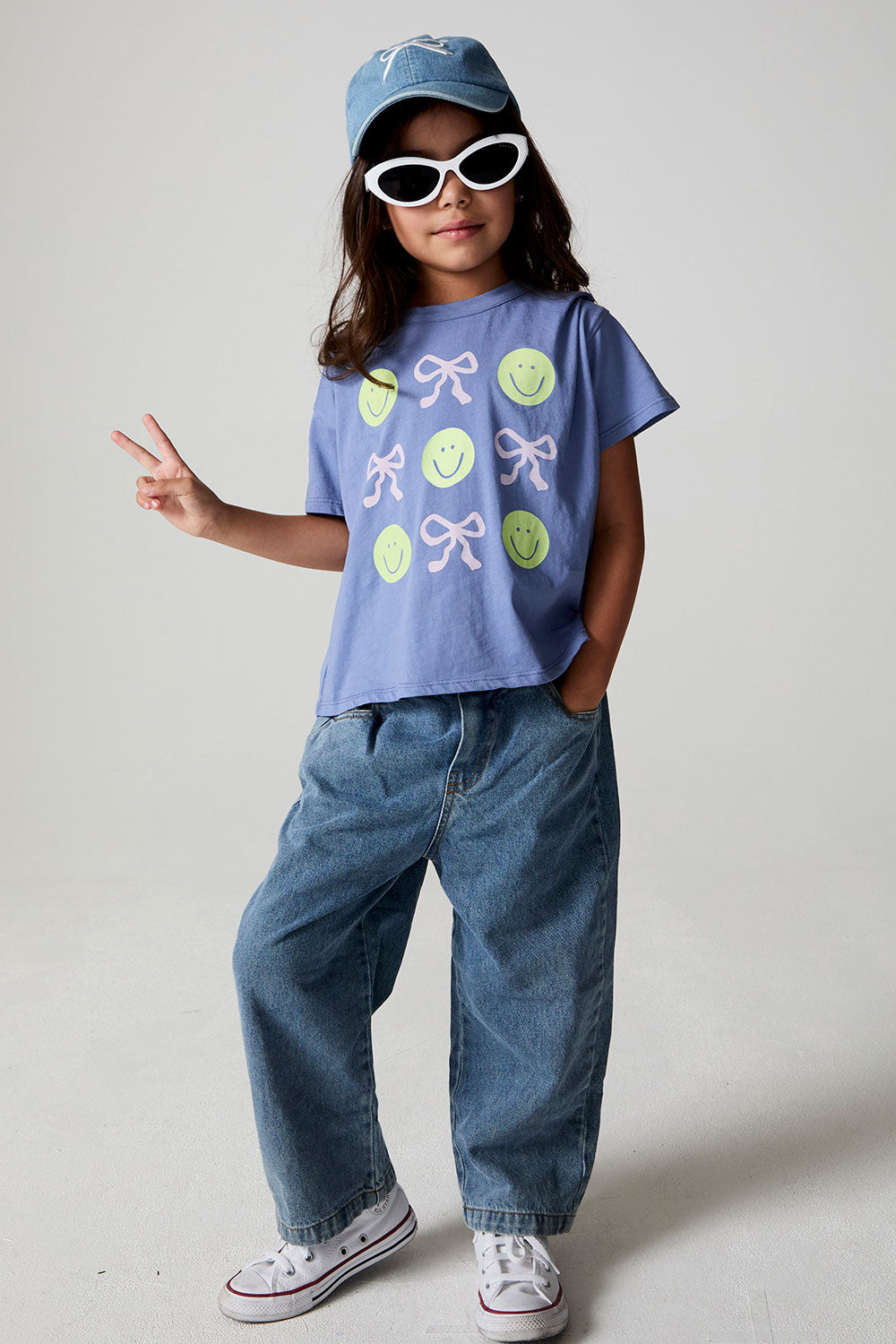 Full body front view of a girl wearing the Chaser Happy Bows Tee with sunglasses and a hat on a neutral background