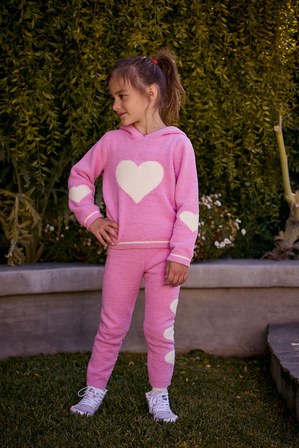 Chaser Kids Holiday Hug Girls Hoodie and Holiday Hug Girls Pants in pink with white hearts, full-body front view outdoors beside a low wall and greenery