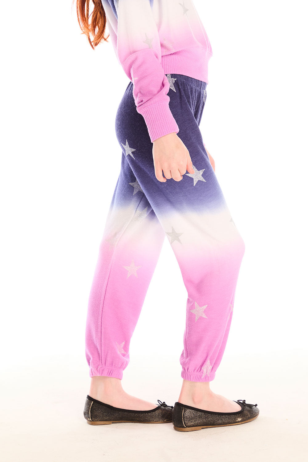 Side view of girl wearing Chaser’s Slim Ombre Tie Dye Pants with colorful glitter star patterns and matching long sleeve top on a white background
