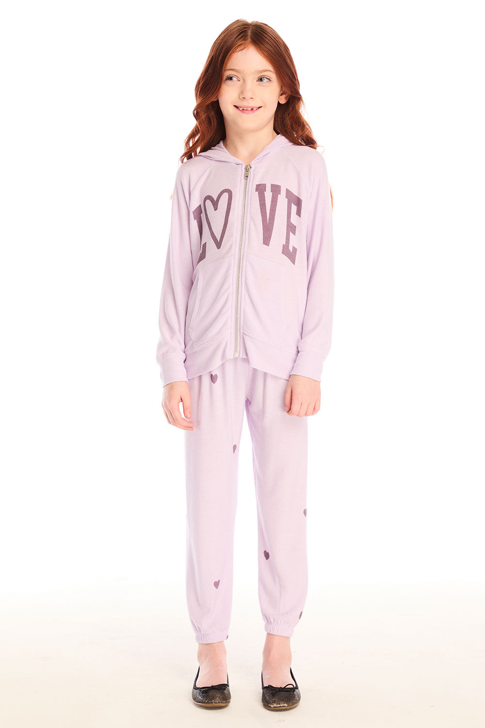 Full body view of girl wearing Chaser’s pink Love Pants with heart prints and matching zip up hoodie with the word “love” across the chest on a white background