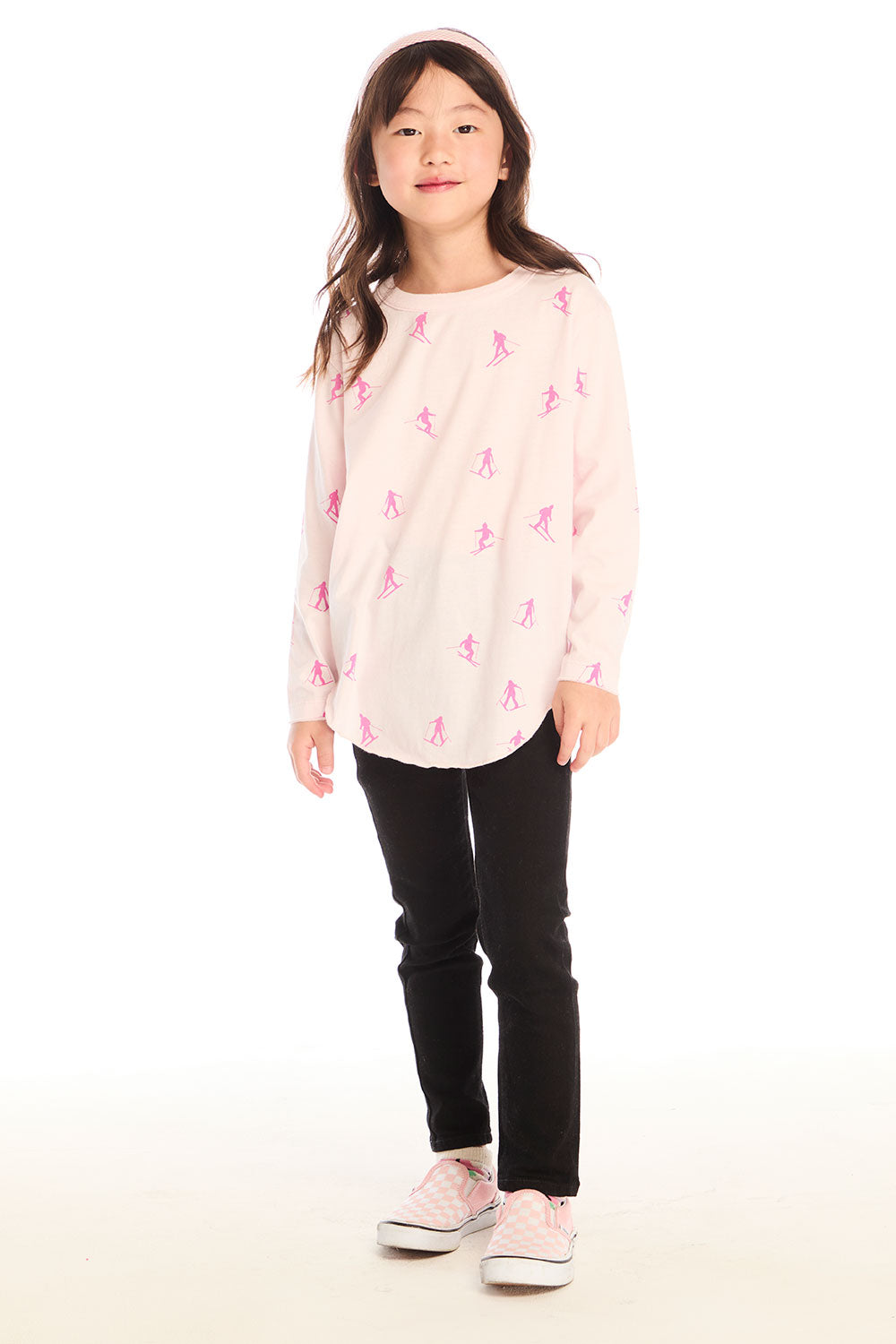 Young girl wearing Chaser's light pink Allover Pink Skiers Long Sleeve top with pink patterns and black pants on a white background