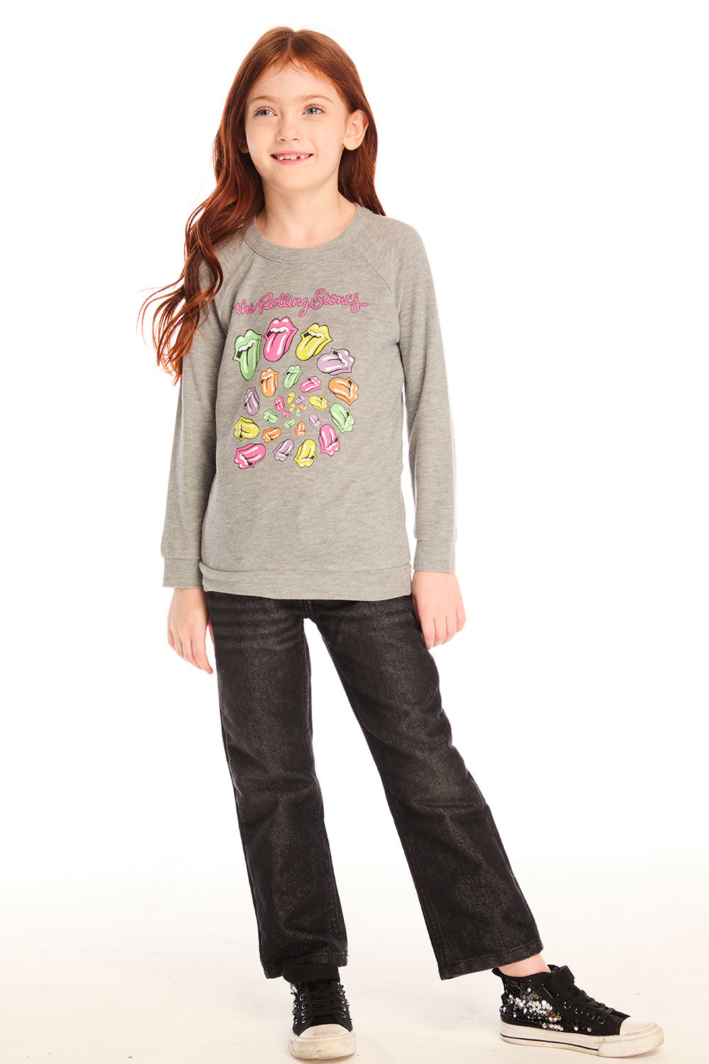 Full body view of girl wearing Chaser’s gray The Rolling Stones Spiral Tongues Long Sleeve with colorful logo designs and text, black jeans, and sparkly shoes on a white background
