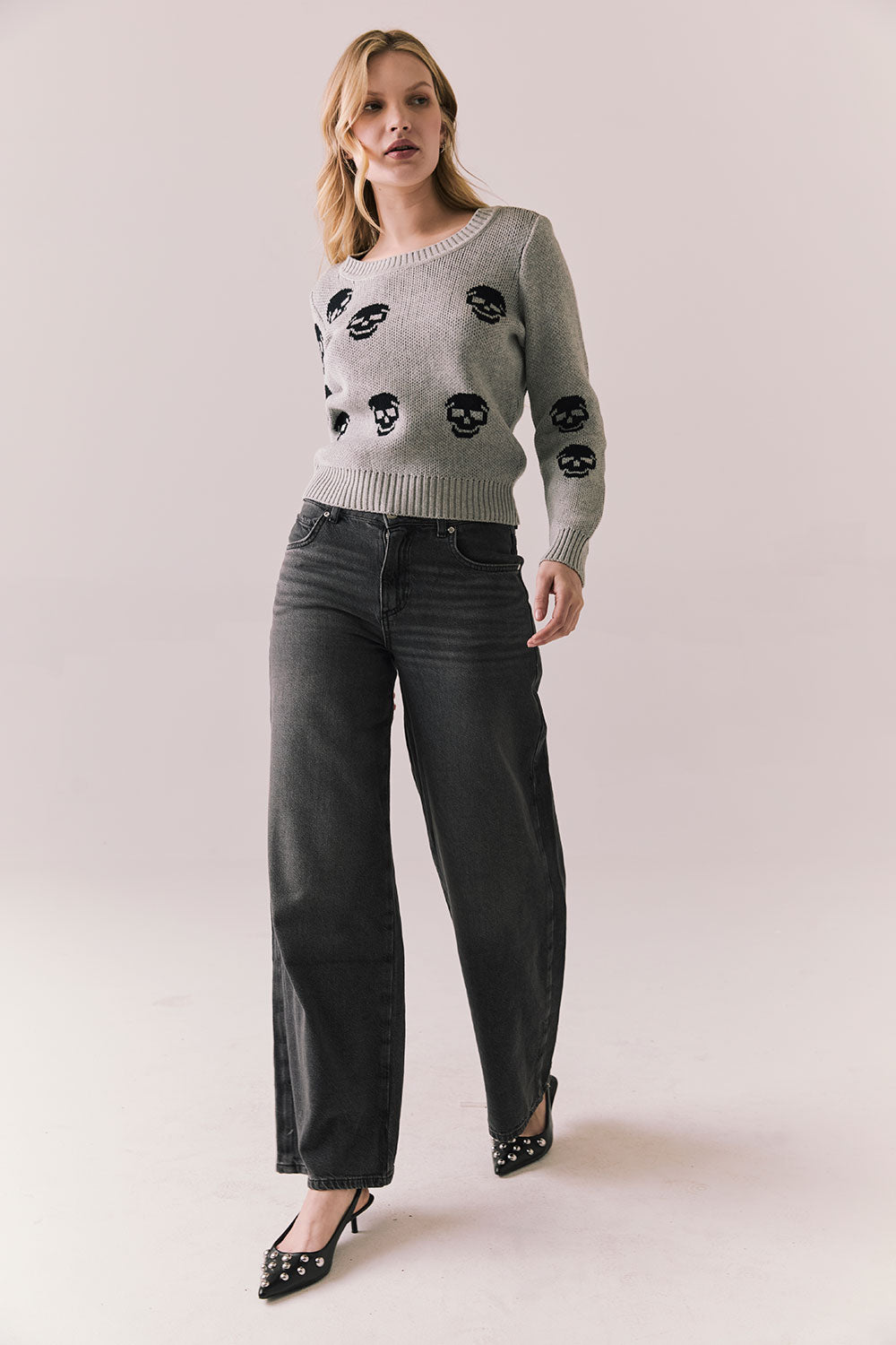 Woman wearing the Tossed Skulls Long Sleeve sweater in Heather Grey with allover black skull motif and fitted silhouette standing against a plain background.