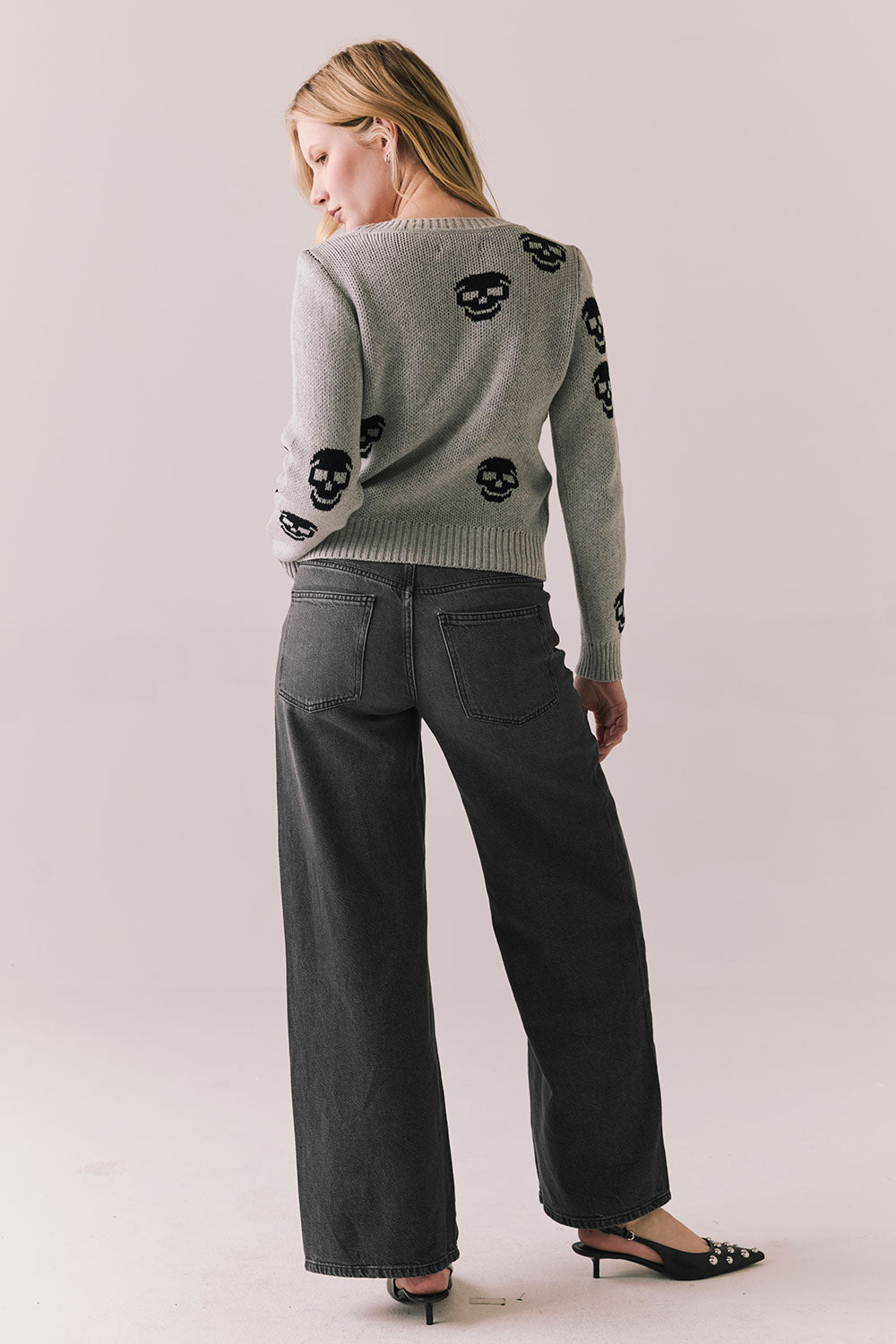 Back view of a woman wearing the Tossed Skulls Long Sleeve sweater in Heather Grey with allover black skull motif and fitted silhouette standing against a plain background.