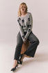 Woman wearing the Tossed Skulls Long Sleeve sweater in Heather Grey with allover black skull motif and fitted silhouette sitting on a wooden block against a plain background.