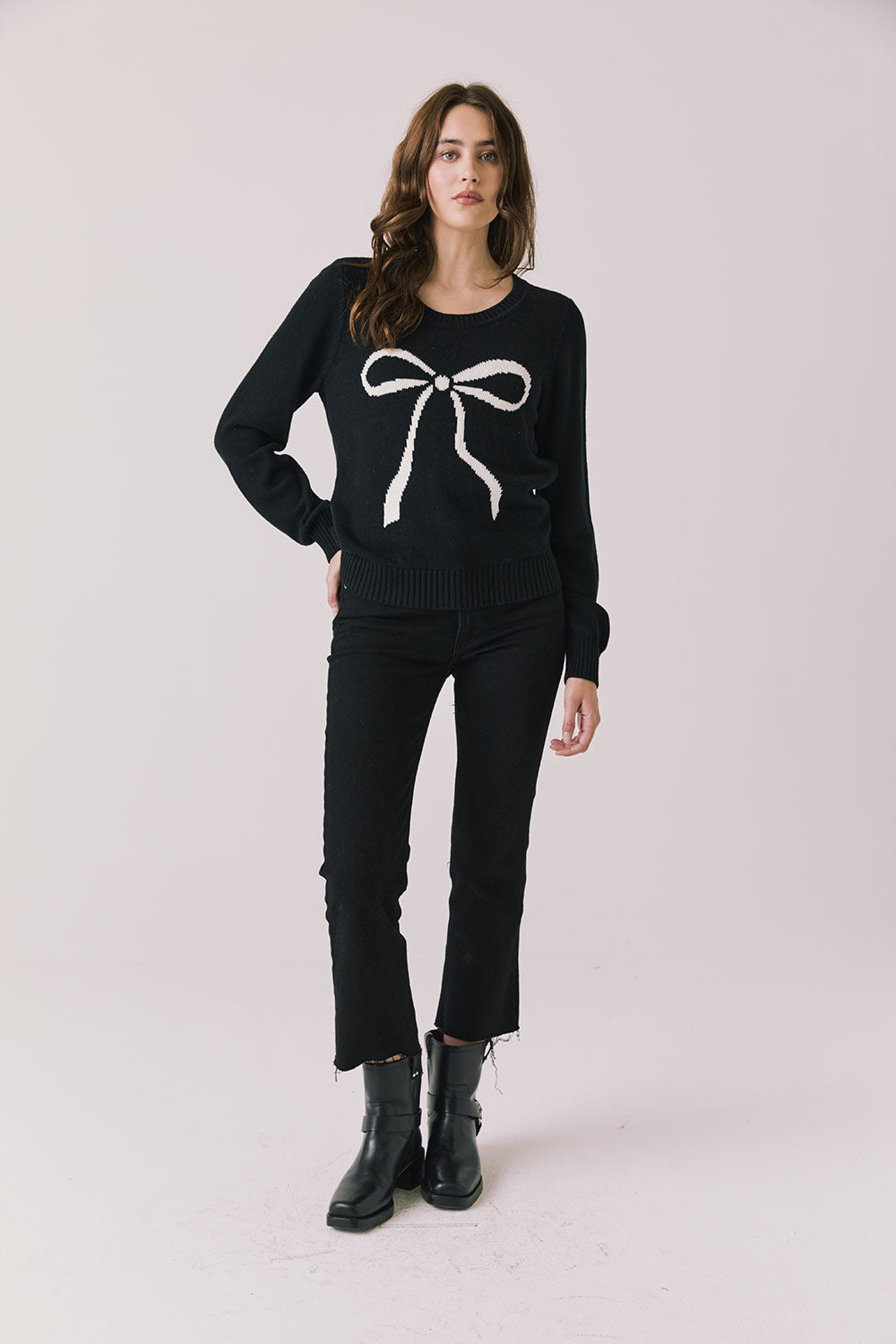 Full body view of woman wearing Chaser’s true black Bow Long Sleeve with white bow design, black jeans, and boots on a plain background.