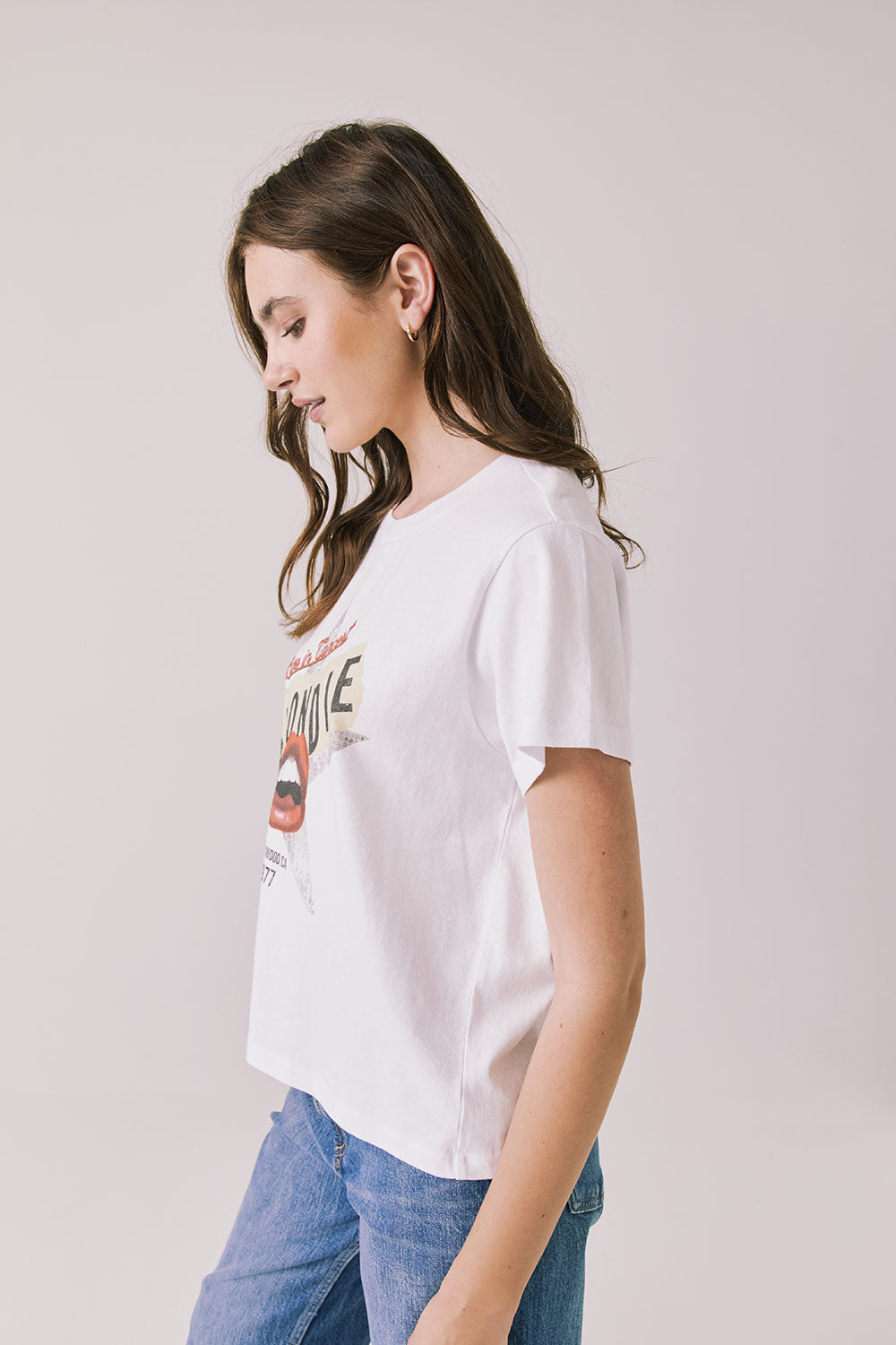 Side view of woman wearing Chaser's white Blondie Live In Hollywood Tee and blue jeans on a plain background