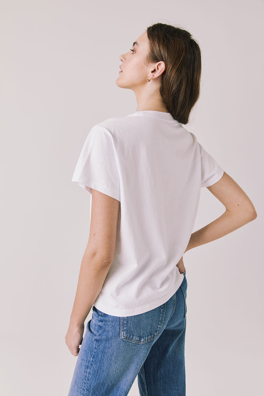 Back view of woman wearing Chaser's white Blondie Live In Hollywood Tee and blue jeans on a plain background