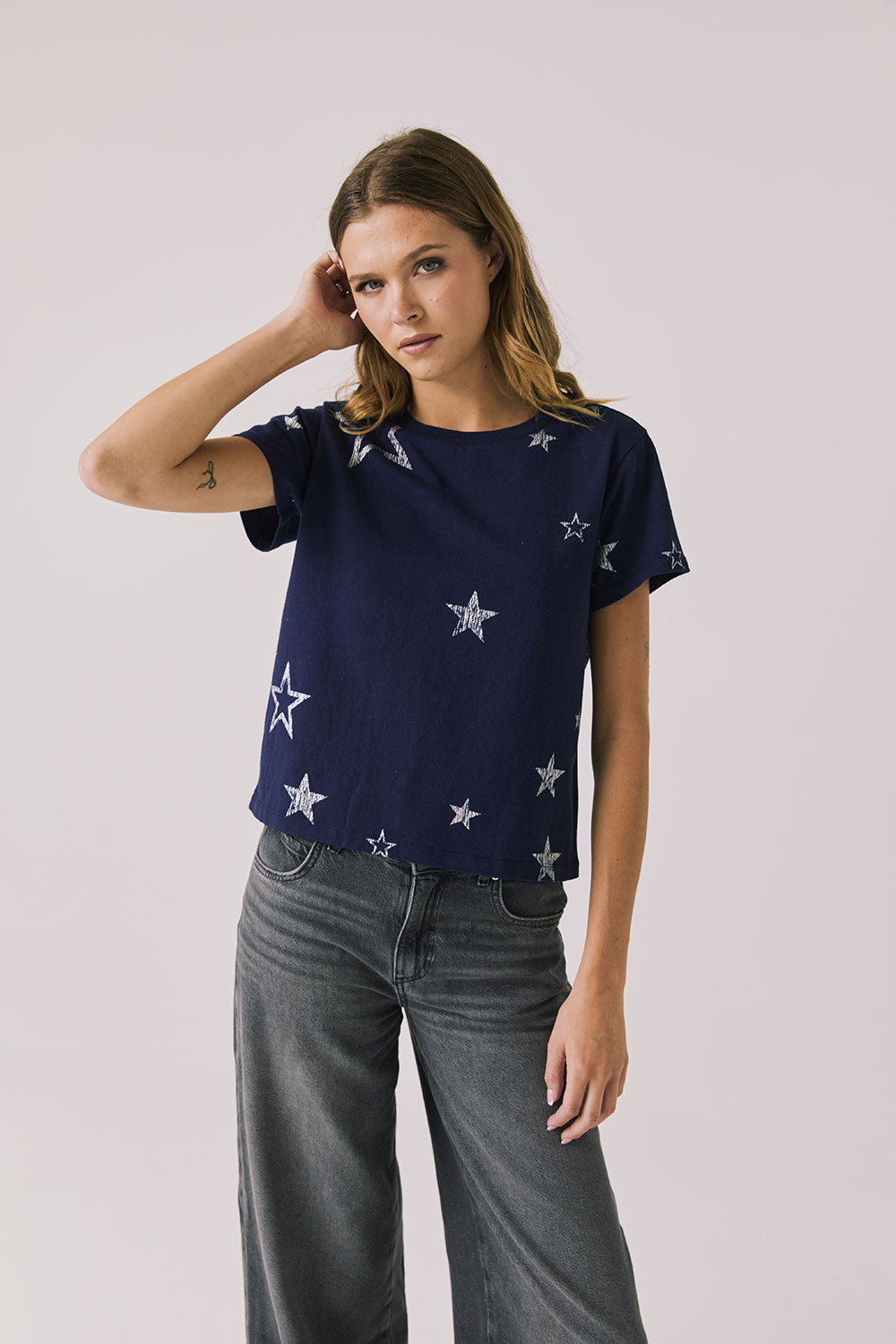 Woman wearing Chaser's Allover Distressed Stars Tee with star patterns and dark denim pants on a plain background