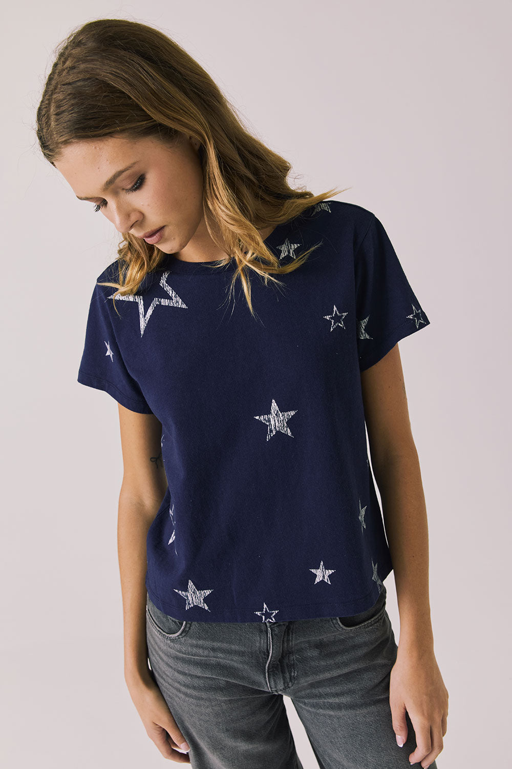 Woman wearing Chaser's Allover Distressed Stars Tee with star patterns and dark denim pants on a plain background