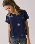 Woman wearing Chaser's Allover Distressed Stars Tee with star patterns and dark denim pants on a plain background