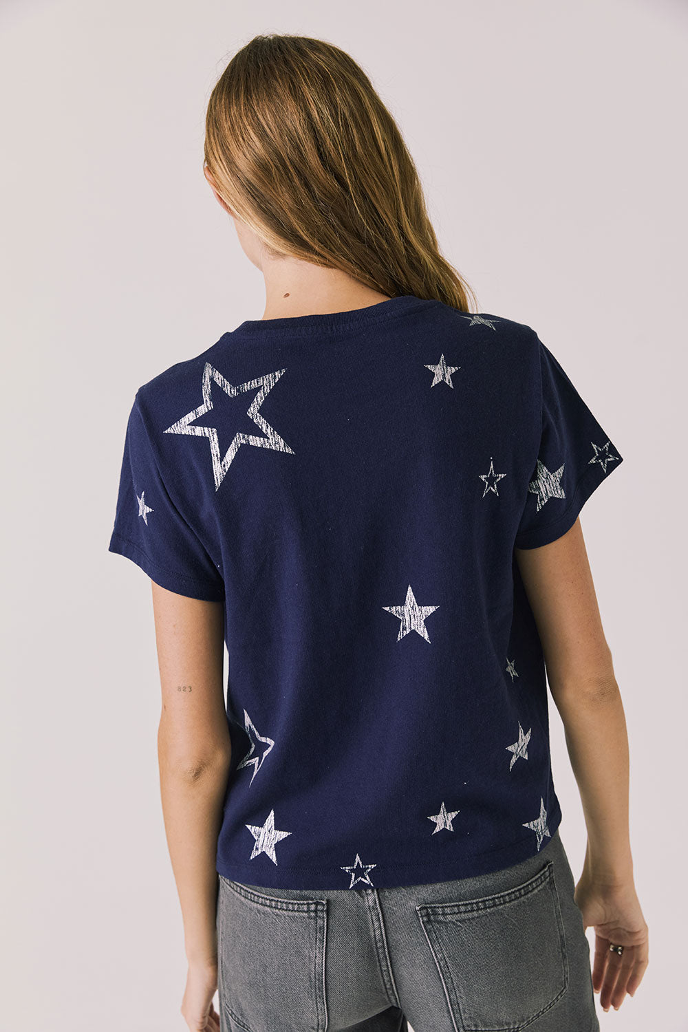 Back view of woman wearing Chaser's Allover Distressed Stars Tee with star patterns and dark denim pants on a plain background
