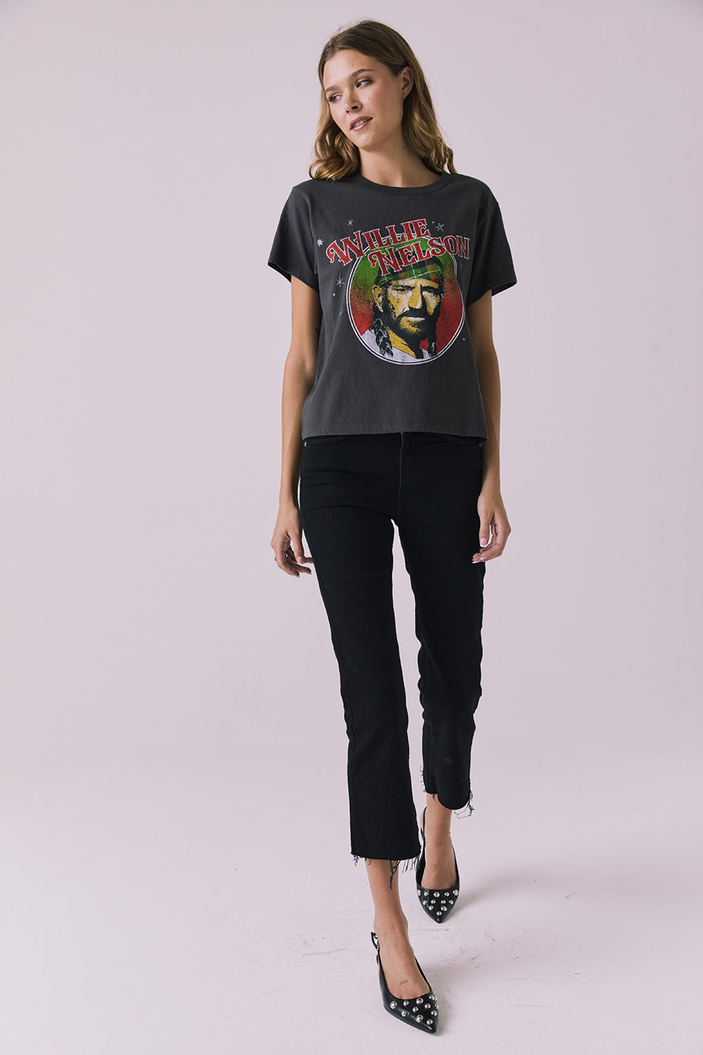Full body view of woman wearing Chaser’s Willie Nelson Willie Christmas Tee with colorful graphic design and blue jeans on a plain background (licensed band collaboration)