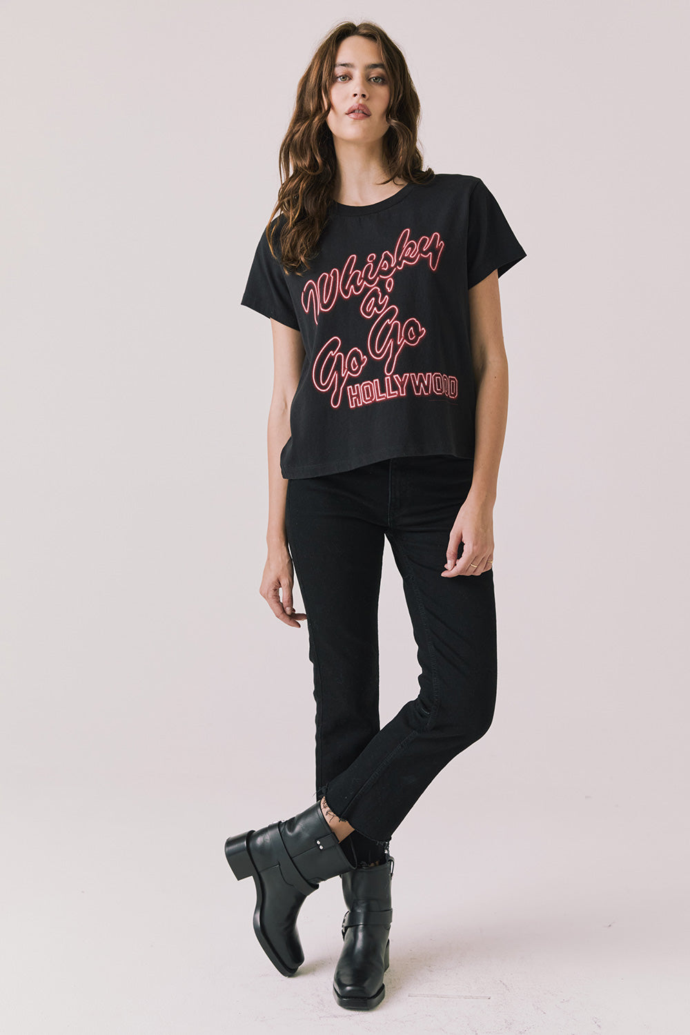 Full body view of woman wearing Chaser’s Whiskey A Go Go Classic Neon Logo black Tee with red text, black pants, and black boots on a light background