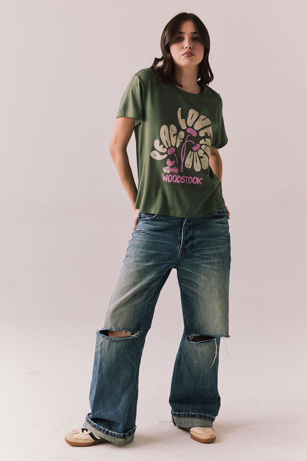 Woman wearing the Woodstock Festival Flower Tee in bronze green with cream and pink floral graphic and vintage font with blue denim against a plain background