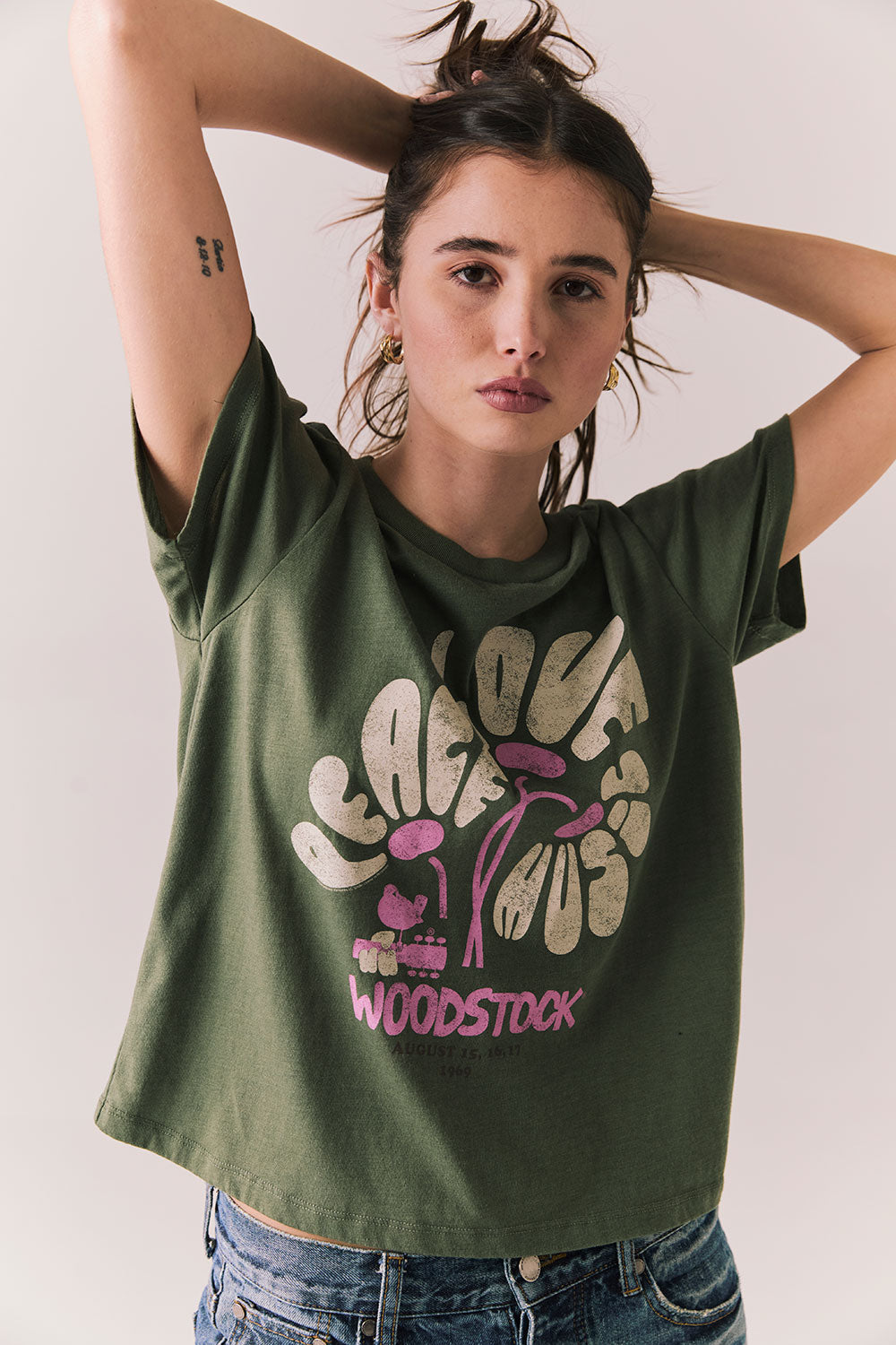 Front view of model wearing Woodstock Festival Flower Tee in bronze green with cream and pink floral graphic and vintage font against a plain background