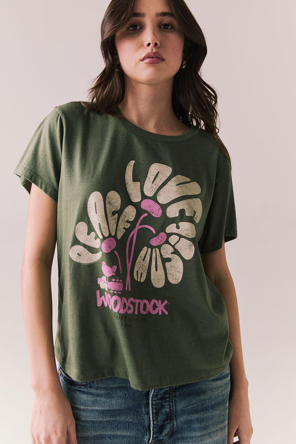 Front view of model wearing Woodstock Festival Flower Tee in bronze green with cream and pink floral graphic and vintage font against a plain background