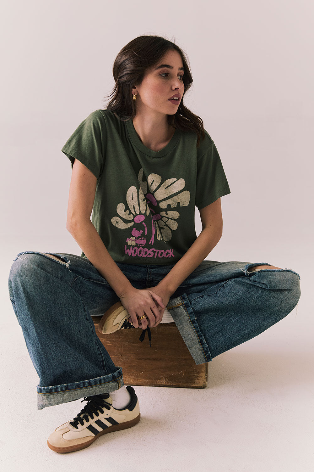Woman wearing the Woodstock Festival Flower Tee in bronze green with cream and pink floral graphic and vintage font with blue denim sitting on a wooden block against a plain background