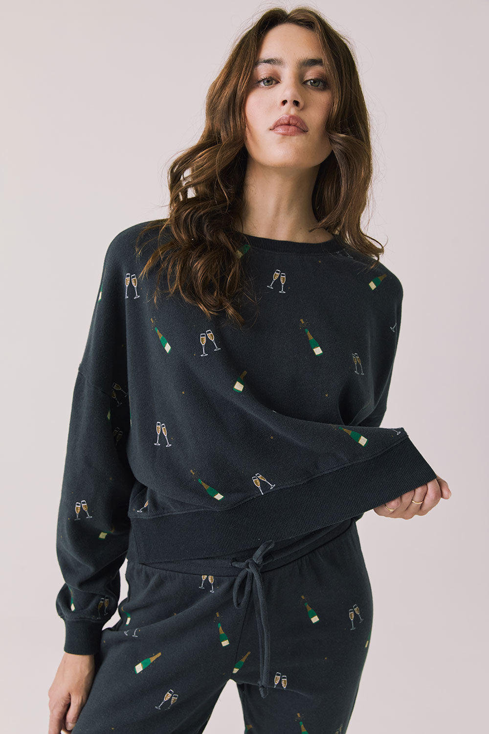 Front view of woman wearing Chaser’s black Champagne Party Long Sleeve Pullover with allover champagne bottle and glass graphics on a light grey background