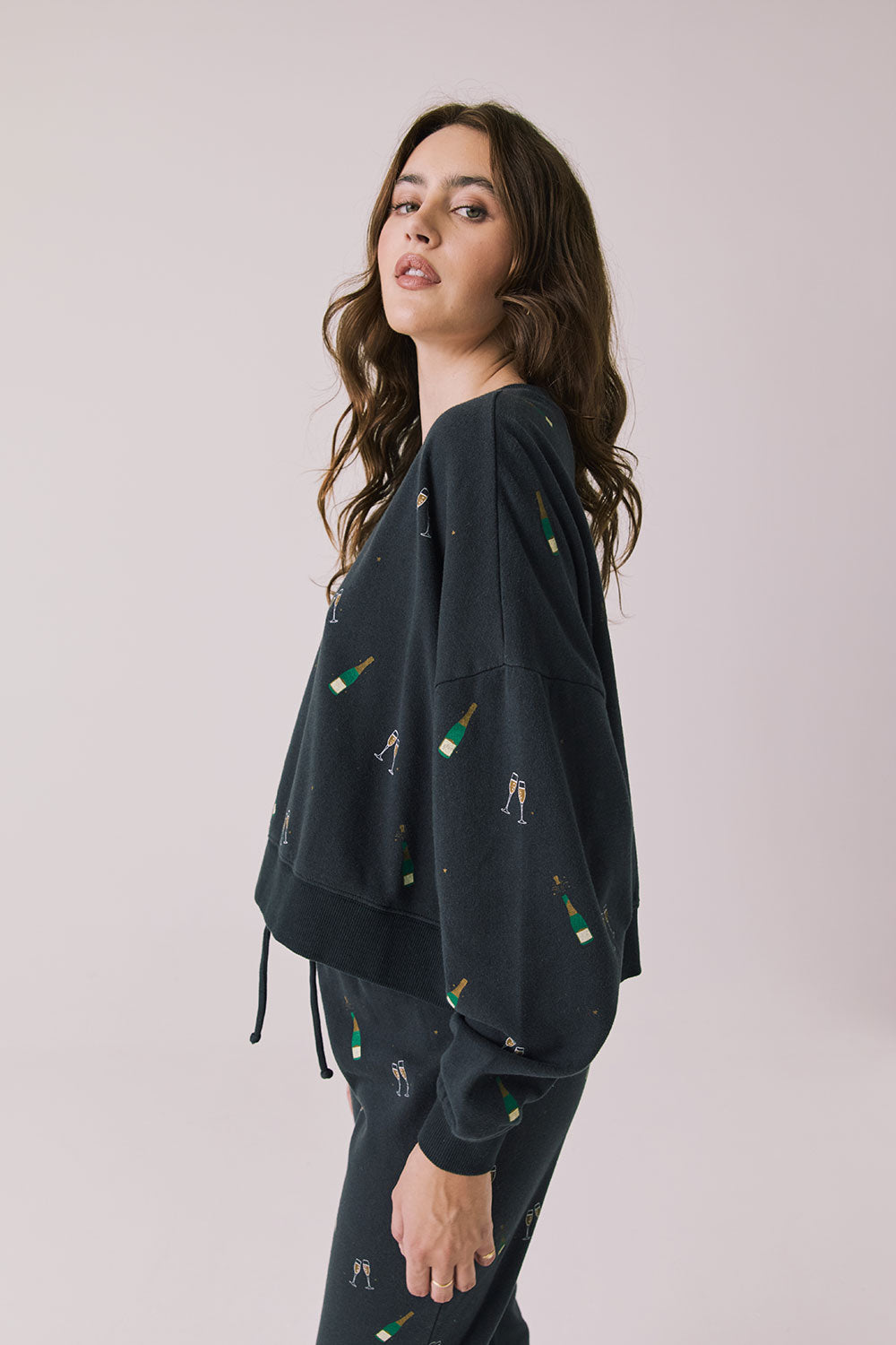 Side view of woman wearing Chaser’s black Champagne Party Long Sleeve Pullover with allover champagne bottle and glass graphics on a light grey background