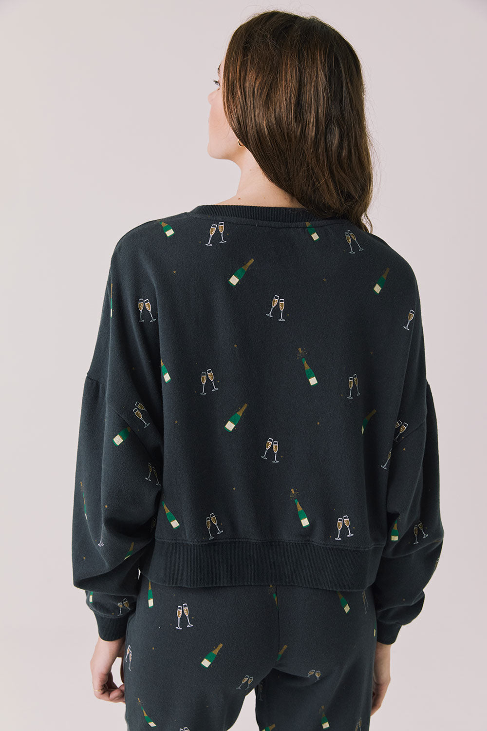 Back view of woman wearing Chaser’s black Champagne Party Long Sleeve Pullover with allover champagne bottle and glass graphics on a light grey background