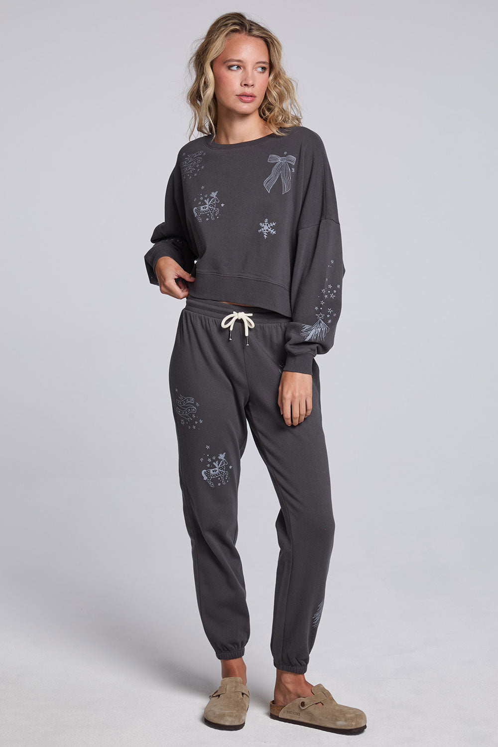 Model wearing Chaser's Holiday Cheer Pullover and matching Holiday Cheer Jogger in Fresh Gravel with scattered holiday icons, full-body view