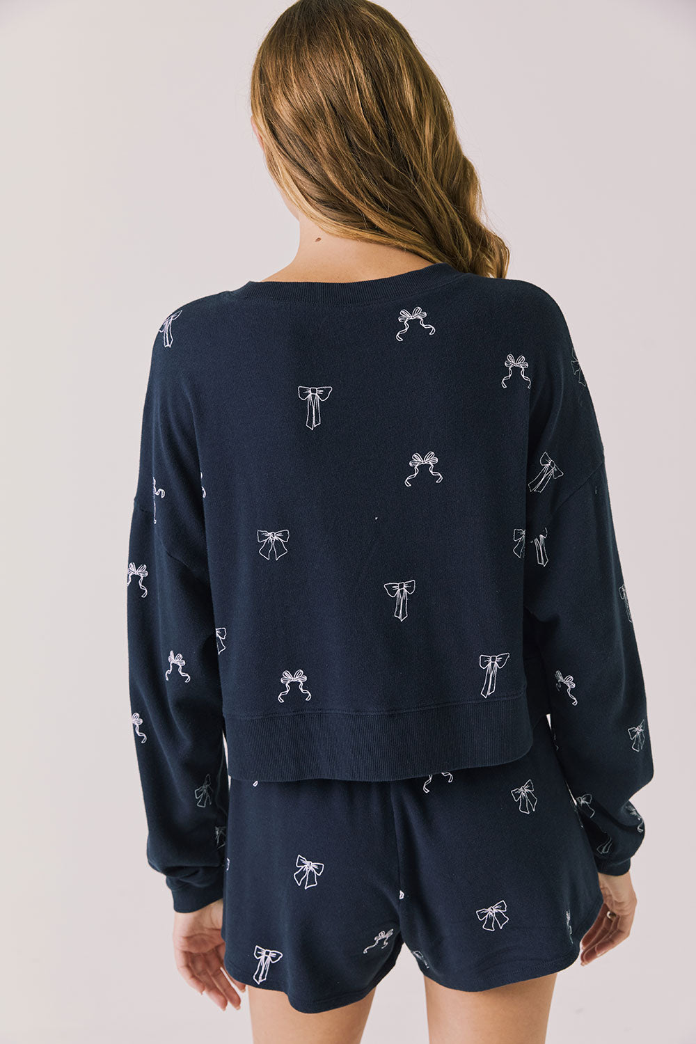 Back view of woman wearing Chaser’s navy Tossed Bows Long Sleeve with allover bow design on a white background