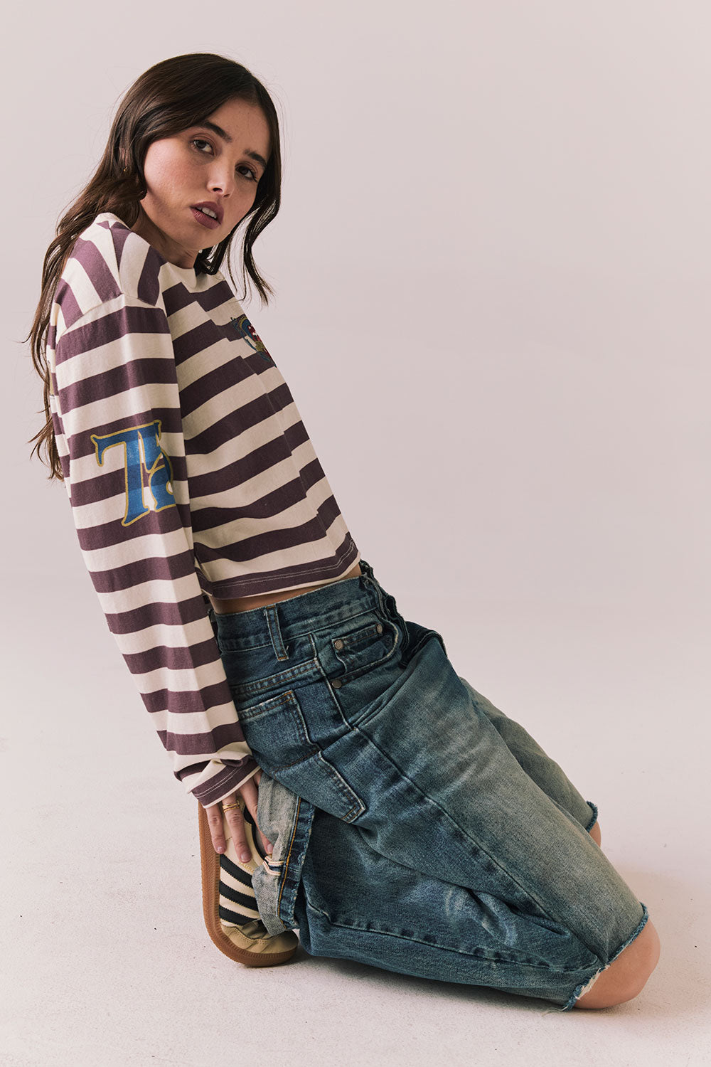 Full side view of model wearingThe Rolling Stones Rugby Stones Logo Long Sleeve with red and white stripes, cropped fit, and tongue logo graphic
and jeans sitting on the floor against a plain background