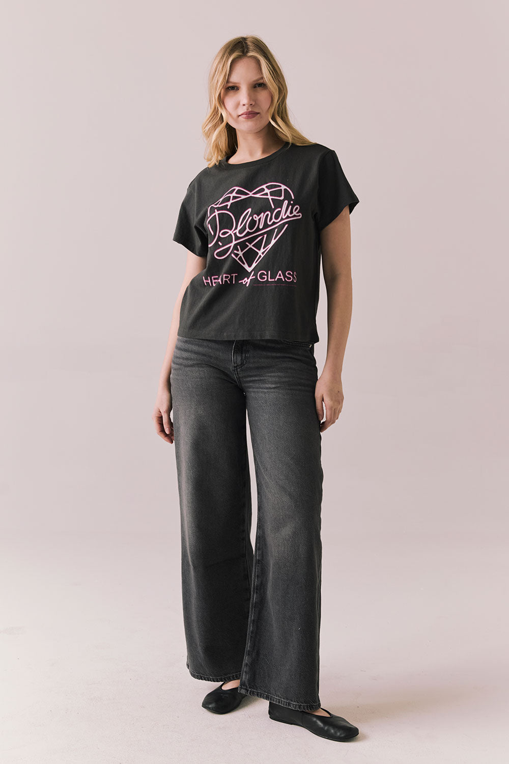 Woman wearing Blondie Heart of Glass tee in black with a graphic design and text, paired with dark jeans on a plain background