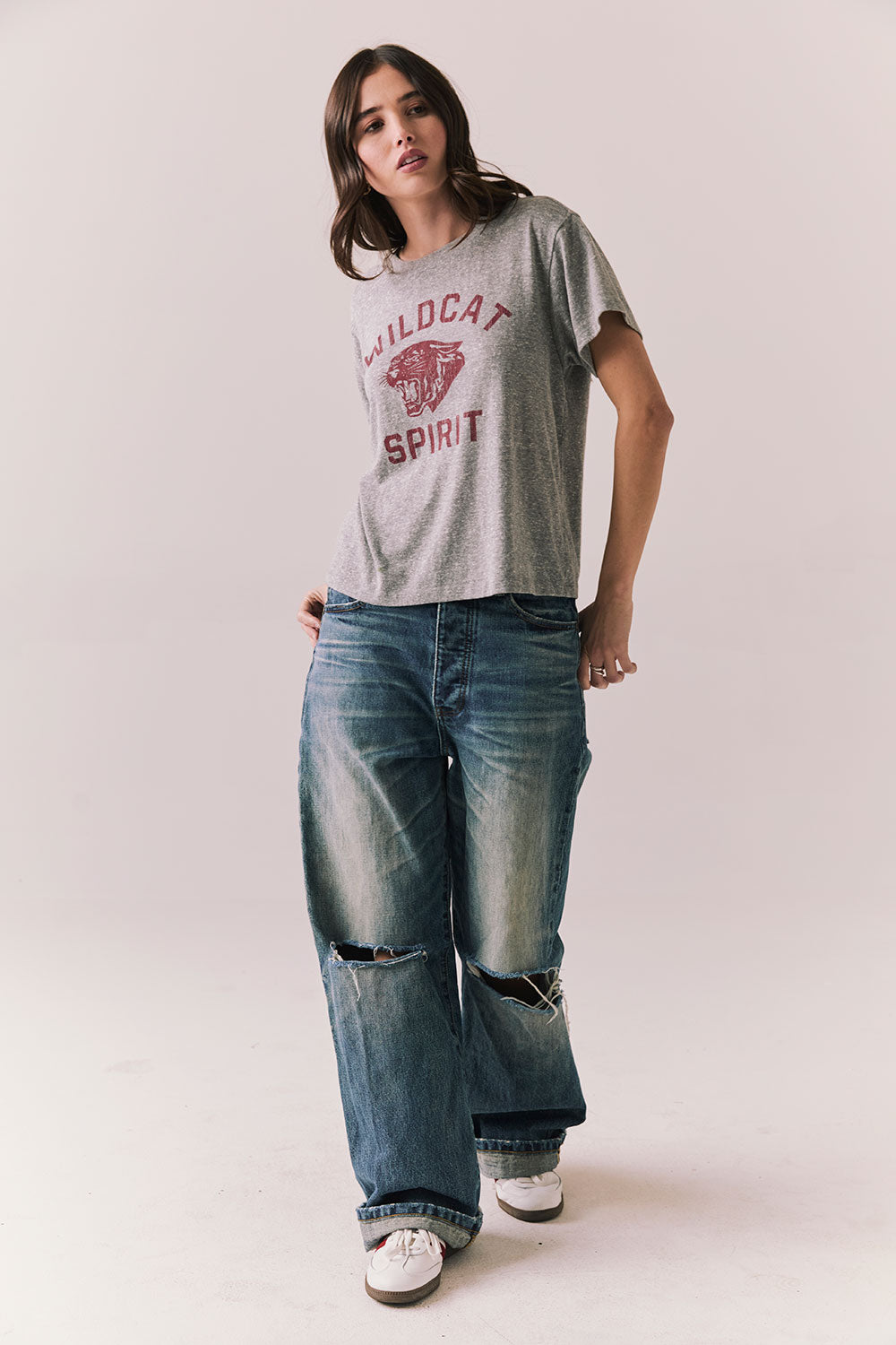 Full front view of model wearing Wildcat Spirit Tee in Heather Grey with maroon wildcat chest graphic with blue jeans, and white sneakers with red stripes on a light background.