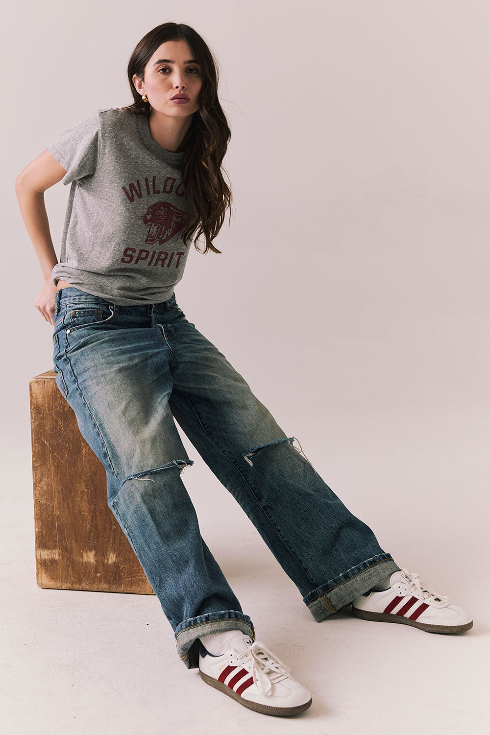 Full front view of model sitting on an apple box wearing Wildcat Spirit Tee in Heather Grey with maroon wildcat chest graphic with blue jeans, and white sneakers with red stripes on a light background.