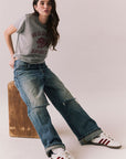 Full front view of model sitting on an apple box wearing Wildcat Spirit Tee in Heather Grey with maroon wildcat chest graphic with blue jeans, and white sneakers with red stripes on a light background.
