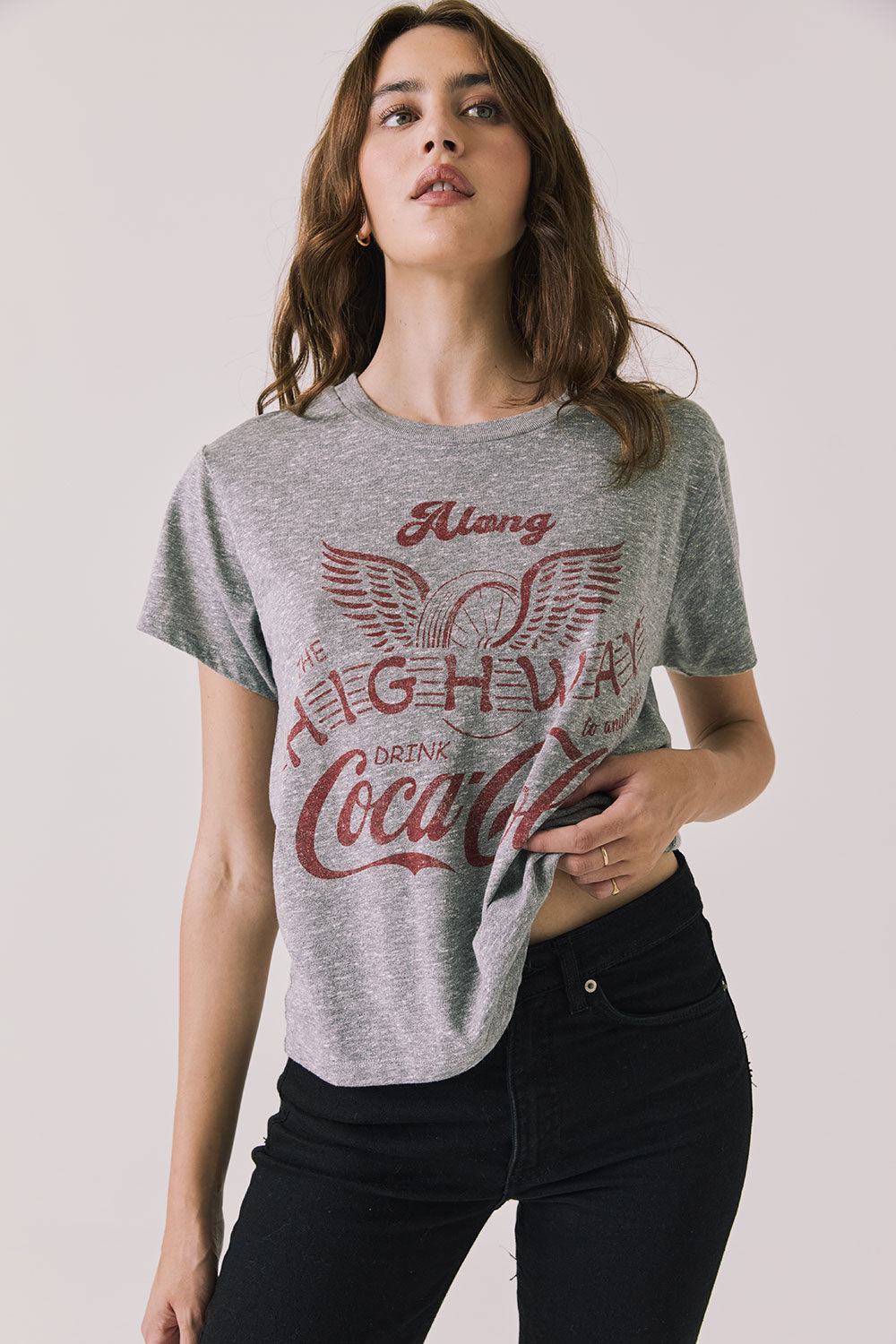 Front view of woman wearing Chaser’s graphic Coca-Cola Highway To Anywhere Tee with black pants on a plain background