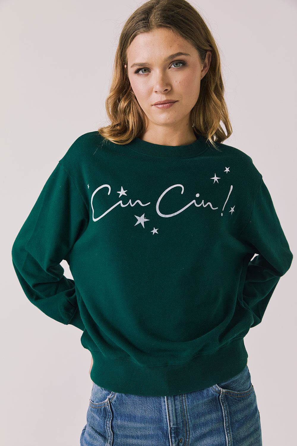 Front view of woman wearing Chaser’s green Cin Cin! Long Sleeve with “Cin Cin” text and star graphics on a plain background