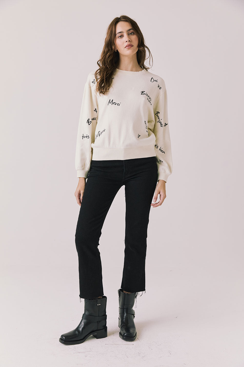 Full body view of woman wearing Chaser’s Starry White French Scripts Long Sleeve with French phrases like “Merci” and “Bonjour” in black text, black pants, and black boots on a white background