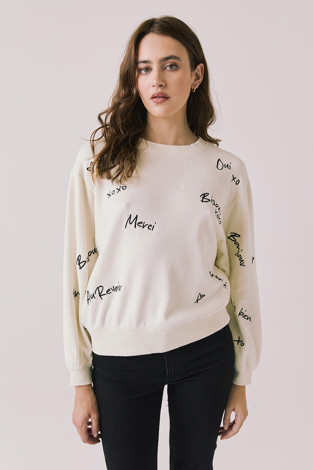 Front view of woman wearing Chaser’s Starry White French Scripts Long Sleeve with French phrases like “Merci” and “Bonjour” in black text on a white background