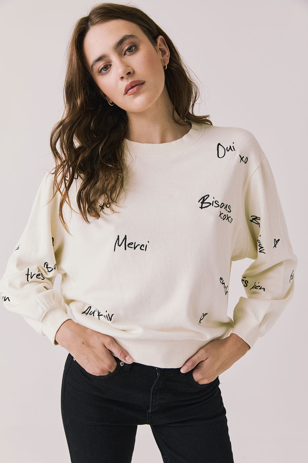 Front view of woman wearing Chaser’s Starry White French Scripts Long Sleeve with French phrases like “Merci” and “Bonjour” in black text on a white background