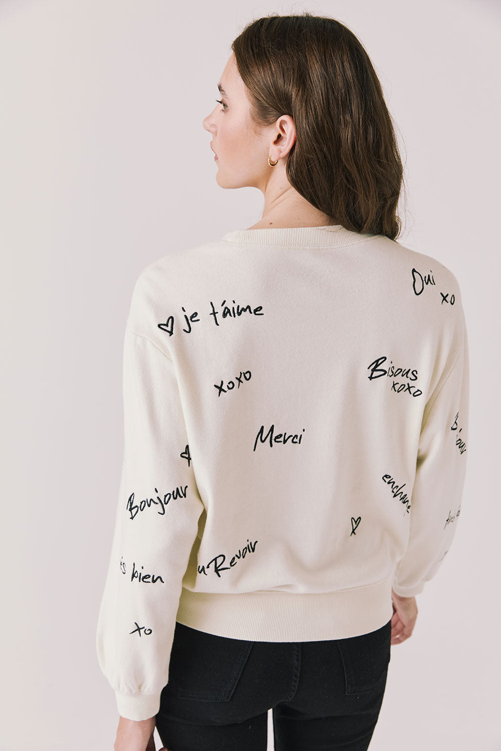 Back view of woman wearing Chaser’s Starry White French Scripts Long Sleeve with French phrases like “Merci” and “Bonjour” in black text on a white background