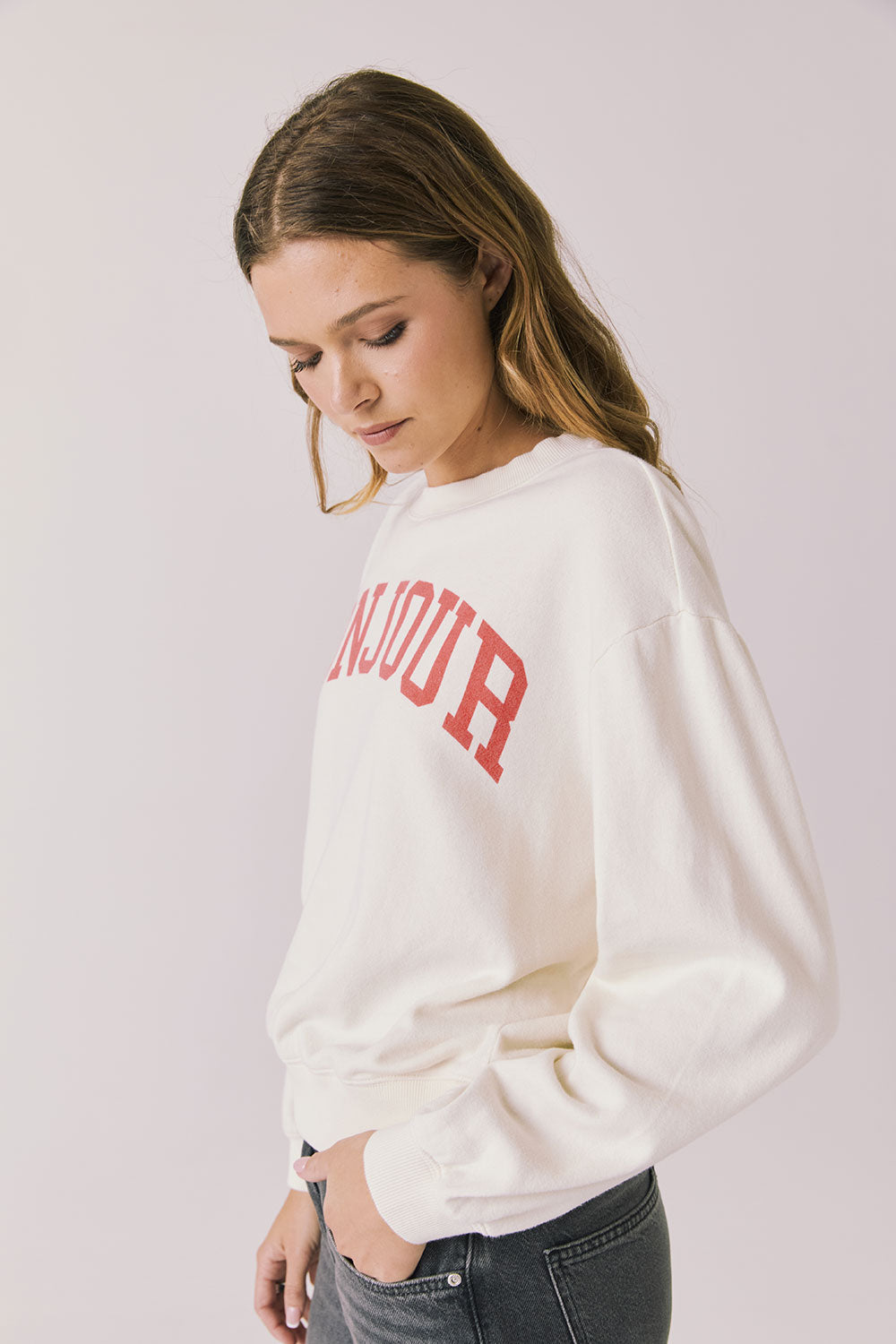 Side view of woman wearing Chaser’s starry white Bonjour Long Sleeve with red ‘Bonjour’ text visible on the front on a plain background.