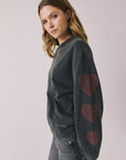 Side view of woman wearing Chaser’s Red Hearts Black Mineral Wash Long Sleeve showing red heart details on the sleeves on a light pink background