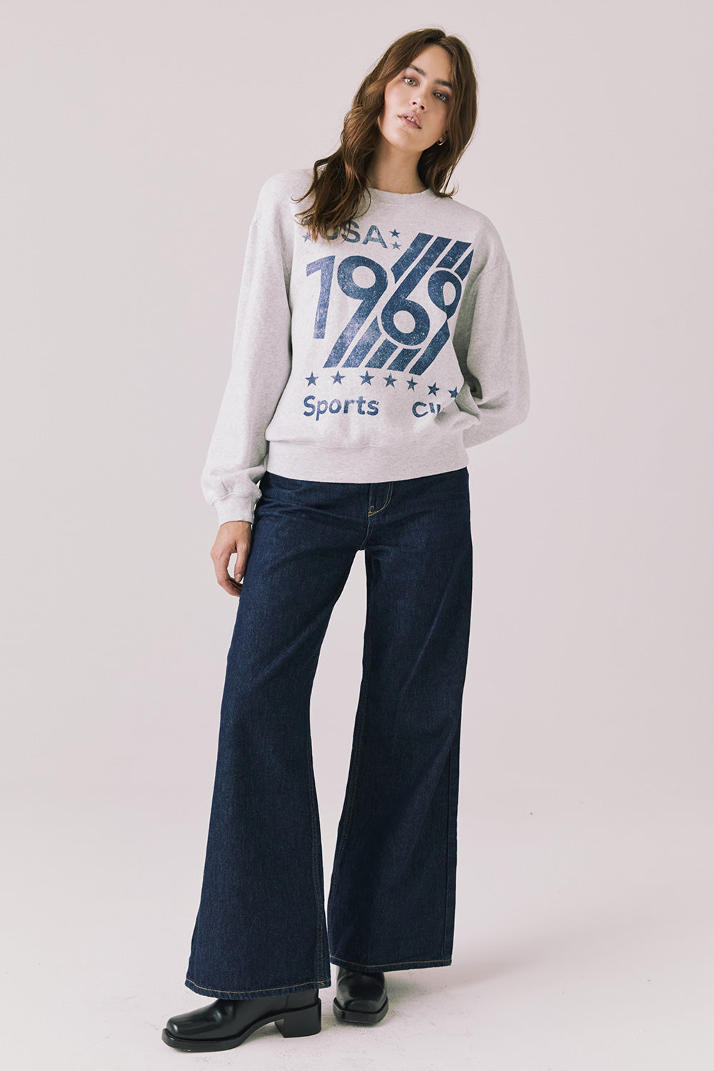 Full body view of woman wearing Chaser’s light grey USA Sports Club Long Sleeve with a blue USA 1969 graphic and dark jeans on a white background
